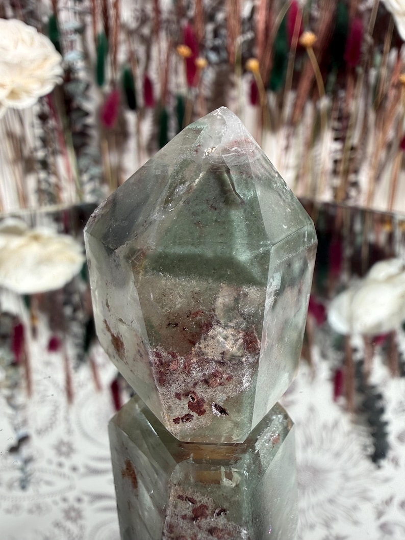 May include: A clear quartz crystal with green and red inclusions. The crystal is faceted with a pointed top and a flat base. The crystal is sitting on a reflective surface. The background is blurred with floral elements.
