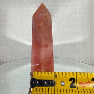 May include: A polished, tower-shaped crystal point with a mottled, translucent coral-pink and white surface. A yellow Milwaukee measuring tape is positioned across the base, indicating a width of approximately 3.2 cm at that point.