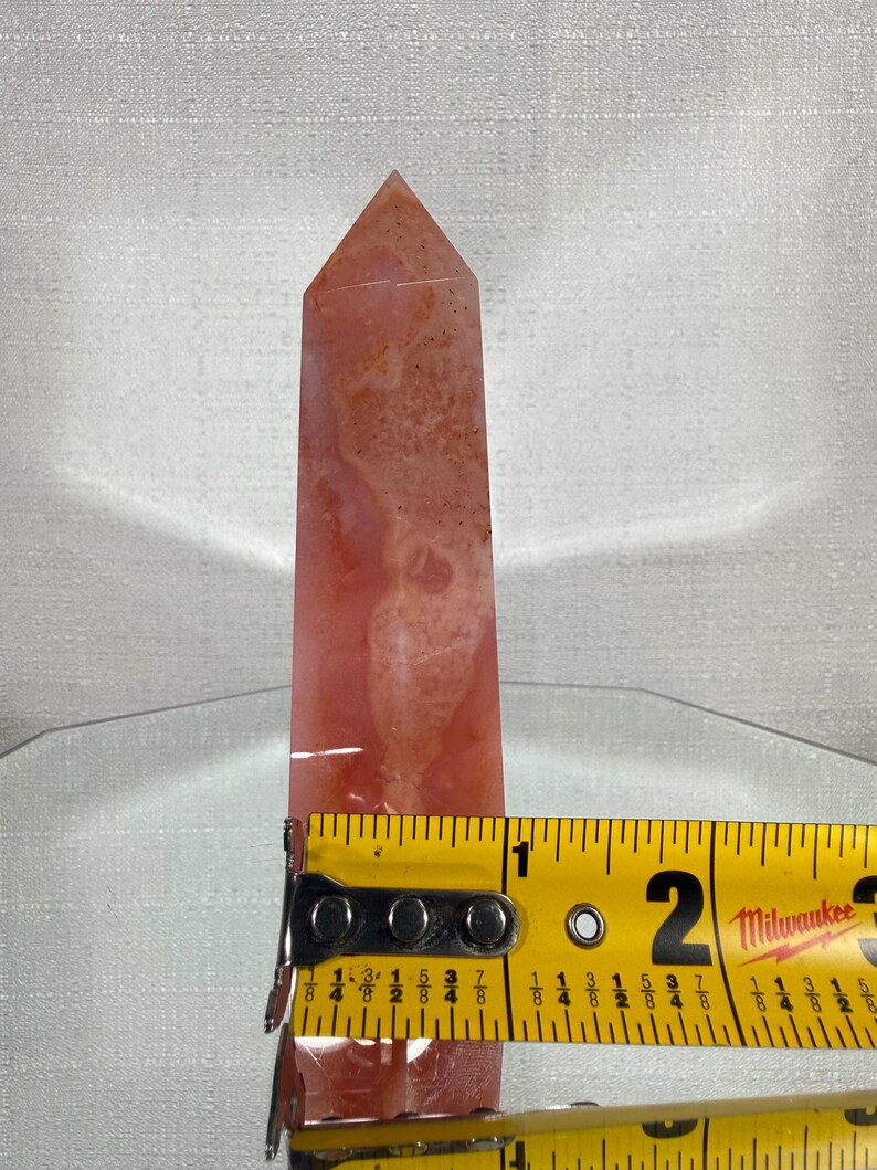 May include: A polished, tower-shaped crystal point with a mottled, translucent coral-pink and white surface. A yellow Milwaukee measuring tape is positioned across the base, indicating a width of approximately 3.2 cm at that point.