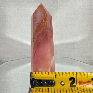 May include: A polished, multi-faceted crystal tower featuring swirling patterns of pink, peach, and translucent white. A yellow Milwaukee measuring tape is positioned across the base, showing a width of approximately 4.45 cm.