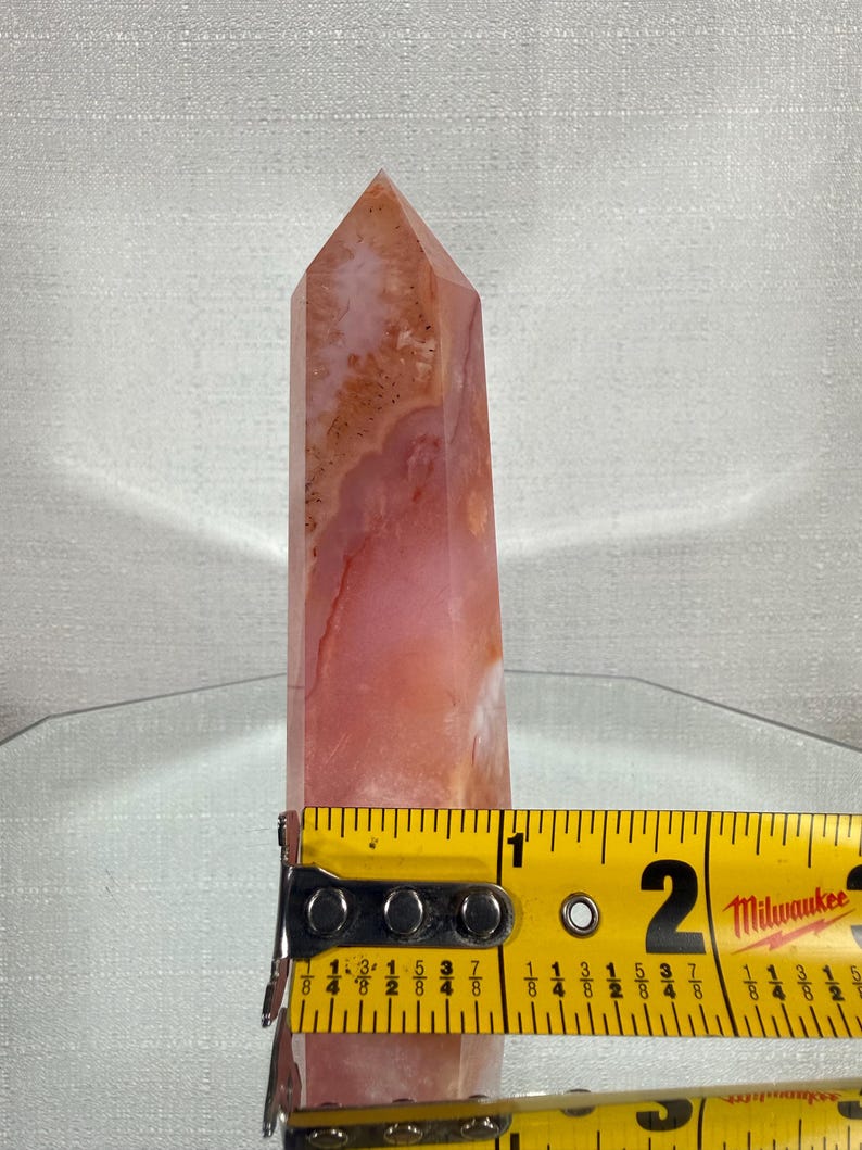 May include: A polished, multi-faceted crystal tower featuring swirling patterns of pink, peach, and translucent white. A yellow Milwaukee measuring tape is positioned across the base, showing a width of approximately 4.45 cm.