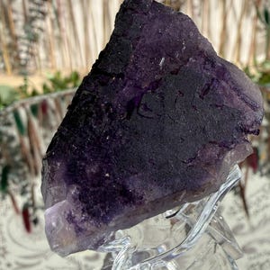 Puede incluir: Cristal de fluorita natural en tonos morados oscuros y negros, exhibido sobre un soporte acr&iacute;lico transparente. El cristal tiene una superficie rugosa y texturizada con diferentes tonos de morado. El fondo est&aacute; borroso, con toques de verde y marr&oacute;n.