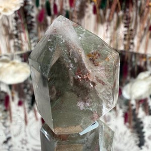 May include: A clear quartz crystal with green and red inclusions. The crystal is cut into a hexagonal shape with a pointed top. The crystal is sitting on a reflective surface, with a blurred background of dried flowers.