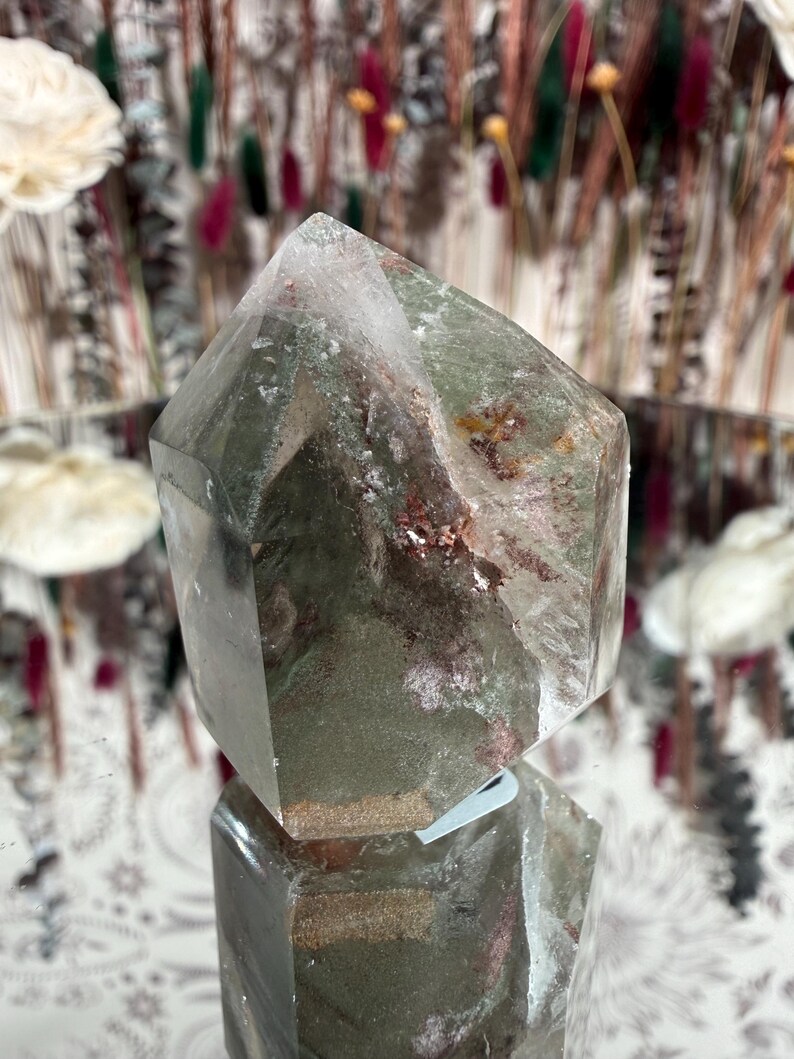 May include: A clear quartz crystal with green and red inclusions. The crystal is cut into a hexagonal shape with a pointed top. The crystal is sitting on a reflective surface, with a blurred background of dried flowers.