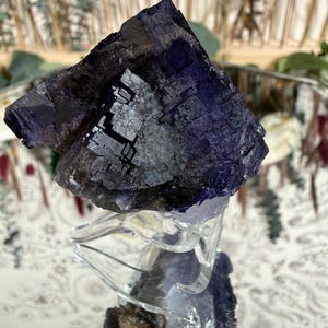 Puede incluir: Un grupo de cristales de fluorita de color p&uacute;rpura oscuro sobre un soporte de vidrio transparente. La superficie del cristal refleja la luz, mostrando su color y textura. El fondo est&aacute; borroso, con toques de vegetaci&oacute;n y un espejo decorativo.