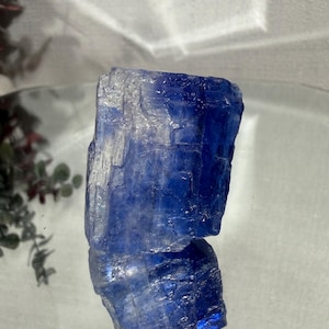 May include: A raw, natural blue kyanite crystal. The crystal has a rough, textured surface with shades of deep blue and lighter, almost white, areas. The crystal is displayed on a reflective surface, creating a mirrored effect.