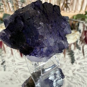 Puede incluir: Un gran grupo de cristales de fluorita de color p&uacute;rpura oscuro con una estructura c&uacute;bica. El cristal est&aacute; montado sobre un soporte acr&iacute;lico transparente, mostrando su forma natural y variaciones de color. La superficie del cristal tiene un aspecto texturizado, con zonas de violeta intenso y negro.
