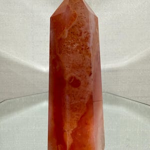 May include: A polished, six-sided crystal tower featuring a warm, mottled blend of orange, coral, and reddish-brown hues. The stone has a smooth, semi-translucent surface with natural internal inclusions and a pointed, faceted top apex.
