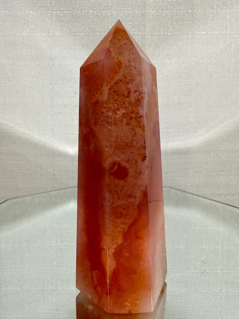 May include: A polished, six-sided crystal tower featuring a warm, mottled blend of orange, coral, and reddish-brown hues. The stone has a smooth, semi-translucent surface with natural internal inclusions and a pointed, faceted top apex.