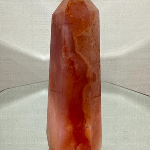 May include: A polished crystal tower with a hexagonal base and a pointed top. The stone features a translucent appearance with a blend of deep orange, reddish-brown, and pale pink hues, displaying natural internal veining and mineral inclusions.