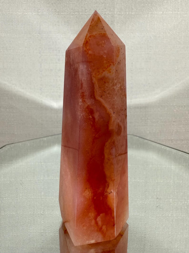 May include: A polished crystal tower with a hexagonal base and a pointed top. The stone features a translucent appearance with a blend of deep orange, reddish-brown, and pale pink hues, displaying natural internal veining and mineral inclusions.