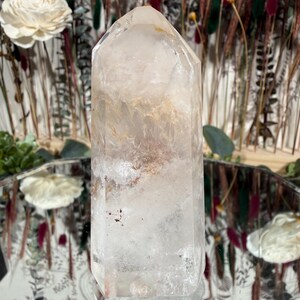 May include: A clear quartz crystal point with internal inclusions of brown and reddish hues. The crystal is faceted and stands upright, reflecting light. The background features dried flowers and a decorative mirror.