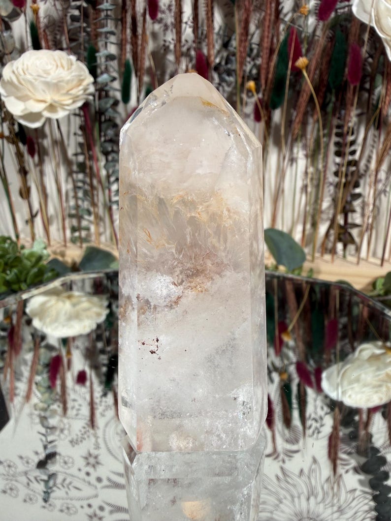 May include: A clear quartz crystal point with internal inclusions of brown and reddish hues. The crystal is faceted and stands upright, reflecting light. The background features dried flowers and a decorative mirror.