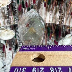 May include: A clear, faceted crystal with internal inclusions, resting near a purple and wooden ruler. The ruler shows measurements in inches. The crystal has a pale green hue and is set against a blurred background of dried flowers and decorative elements.