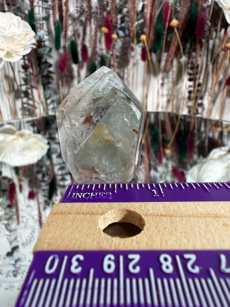 May include: A clear, faceted crystal with internal inclusions, resting near a purple and wooden ruler. The ruler shows measurements in inches. The crystal has a pale green hue and is set against a blurred background of dried flowers and decorative elements.
