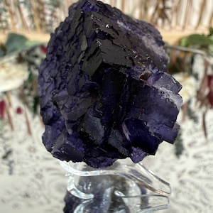 Puede incluir: Un gran cristal de fluorita en bruto, en tonos morados oscuros y negros, exhibido sobre un soporte acr&iacute;lico transparente. La superficie del cristal tiene un aspecto texturizado y geom&eacute;trico, con estructuras cristalinas visibles. El fondo est&aacute; borroso.
