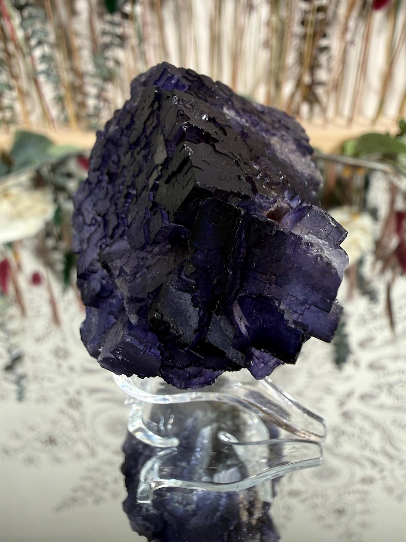 Puede incluir: Un gran cristal de fluorita en bruto, en tonos morados oscuros y negros, exhibido sobre un soporte acr&iacute;lico transparente. La superficie del cristal tiene un aspecto texturizado y geom&eacute;trico, con estructuras cristalinas visibles. El fondo est&aacute; borroso.