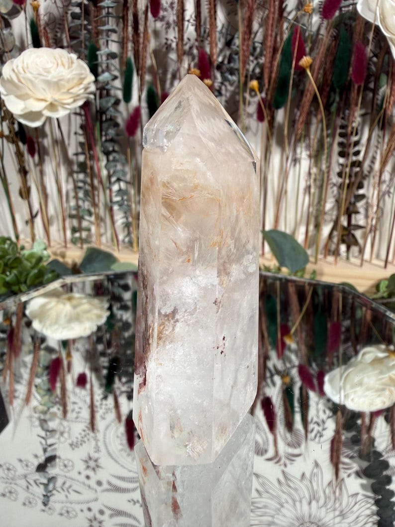 May include: A clear quartz crystal point with internal inclusions and a polished surface. The crystal is standing upright, reflecting in a mirrored surface. The background includes dried flowers and decorative elements.