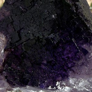 Puede incluir: Primer plano de un cristal de fluorita natural. El cristal presenta un tono p&uacute;rpura intenso con patrones geom&eacute;tricos oscuros, casi negros, en su superficie. La parte inferior del cristal tiene una apariencia m&aacute;s clara y transl&uacute;cida.
