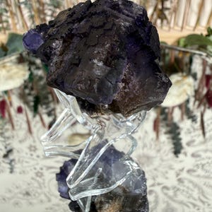 Puede incluir: Un grupo de cristales de fluorita de color p&uacute;rpura oscuro con una superficie rugosa y texturizada. El cristal se asienta sobre un soporte transparente similar al vidrio, reflejando su color y forma. El color del cristal var&iacute;a del p&uacute;rpura oscuro a tonos m&aacute;s claros.