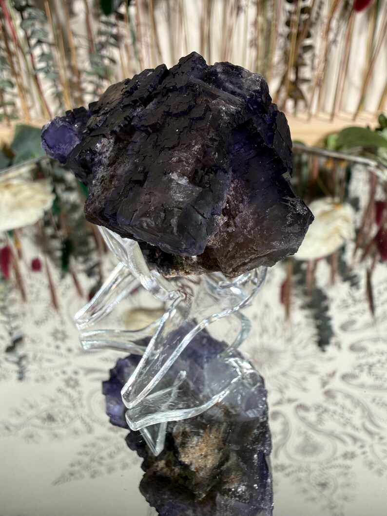 Puede incluir: Un grupo de cristales de fluorita de color p&uacute;rpura oscuro con una superficie rugosa y texturizada. El cristal se asienta sobre un soporte transparente similar al vidrio, reflejando su color y forma. El color del cristal var&iacute;a del p&uacute;rpura oscuro a tonos m&aacute;s claros.