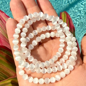 May include: A strand of white, round gemstone beads, likely selenite, is held in a hand. The beads have a milky, translucent appearance. The background features a blue water surface and a hint of tropical foliage.