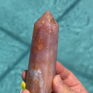 May include: A polished, six-sided crystal tower featuring a translucent, dusty rose and mauve colour palette. The stone displays internal cloudy white inclusions and warm, amber-toned patches throughout its vertical, faceted structure.
