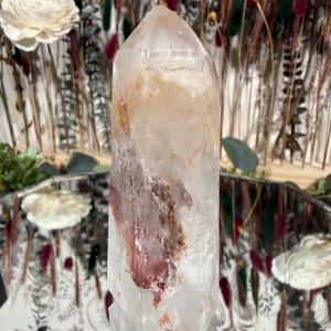 May include: A clear quartz crystal point with reddish-brown inclusions. The crystal is faceted and stands upright, reflecting light. The background includes dried flowers and a mirror, suggesting a natural or spiritual theme.