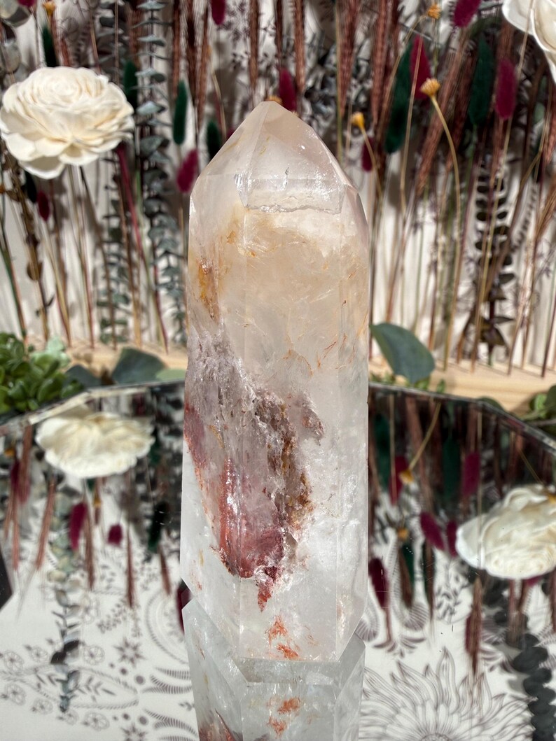 May include: A clear quartz crystal point with reddish-brown inclusions. The crystal is faceted and stands upright, reflecting light. The background includes dried flowers and a mirror, suggesting a natural or spiritual theme.