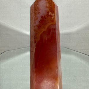 May include: A polished crystal tower with a hexagonal point, featuring a vibrant blend of deep orange, reddish-brown, and translucent white mineral inclusions. The stone displays natural banding and internal crystalline textures throughout its surface.