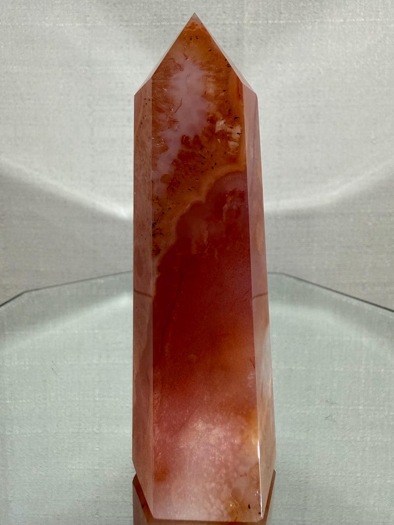 May include: A polished crystal tower with a hexagonal point, featuring a vibrant blend of deep orange, reddish-brown, and translucent white mineral inclusions. The stone displays natural banding and internal crystalline textures throughout its surface.