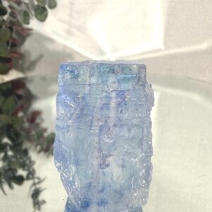 May include: A light blue, raw crystal with a rough, textured surface. The crystal is a rectangular shape and reflects light, creating a shimmering effect. The crystal is a natural gemstone.