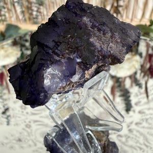 Puede incluir: Un esp&eacute;cimen de cristal de fluorita p&uacute;rpura oscuro con una superficie en capas y texturizada. El cristal est&aacute; montado sobre un soporte transparente y geom&eacute;trico, mostrando su belleza natural. La fluorita tiene un color profundo y rico.