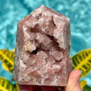 May include: A dusty rose-colored crystal tower featuring a central geode cavity filled with small, sparkling crystalline structures. The mineral specimen has a pointed top and a rough, textured surface with varying shades of pink and light brown.