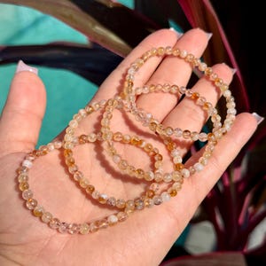 May include: Four delicate bracelets made of small, round, amber-colored beads. The bracelets are displayed on a hand, with a background of green and red foliage. The beads have a translucent quality, and the bracelets appear to be strung on elastic.