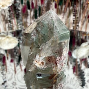 May include: A clear quartz crystal point with internal green and reddish inclusions. The crystal is faceted and reflects light, showcasing its natural beauty. The crystal is displayed on a reflective surface.