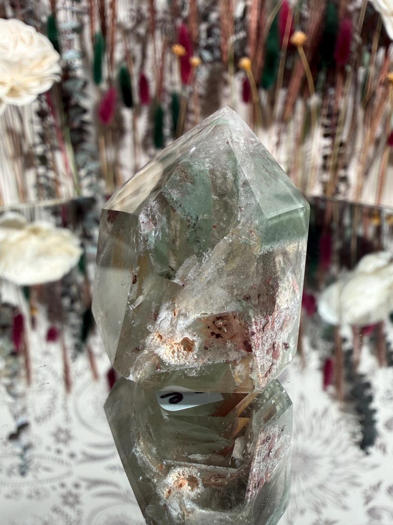May include: A clear quartz crystal point with internal green and reddish inclusions. The crystal is faceted and reflects light, showcasing its natural beauty. The crystal is displayed on a reflective surface.