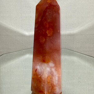 May include: A polished, six-sided crystal tower featuring a blend of translucent orange, deep red, and opaque white mineral inclusions. The stone has a smooth, faceted surface that tapers to a point at the top, displaying natural colour variations.