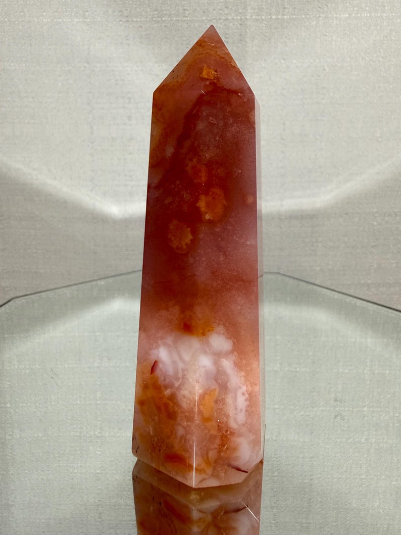May include: A polished, six-sided crystal tower featuring a blend of translucent orange, deep red, and opaque white mineral inclusions. The stone has a smooth, faceted surface that tapers to a point at the top, displaying natural colour variations.