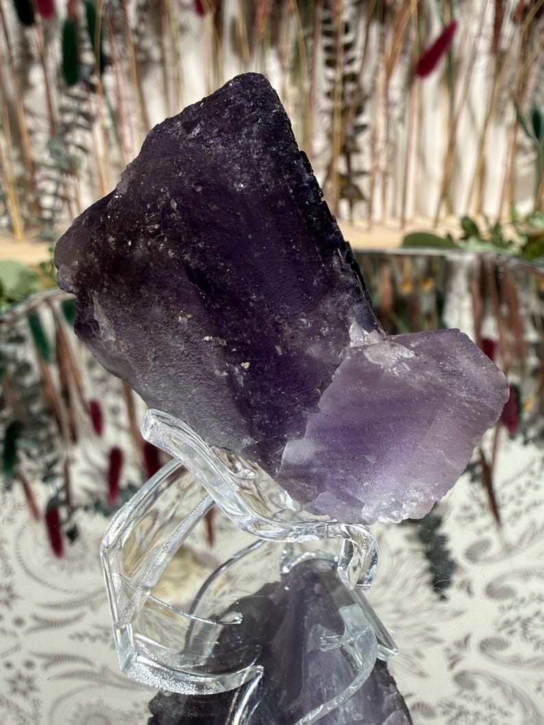 Puede incluir: Un gran cristal de amatista en bruto, en tonos morados profundos y lavanda, se exhibe sobre un soporte decorativo transparente. El cristal tiene una superficie rugosa y texturizada y est&aacute; iluminado por luz natural. El fondo presenta plantas secas borrosas.