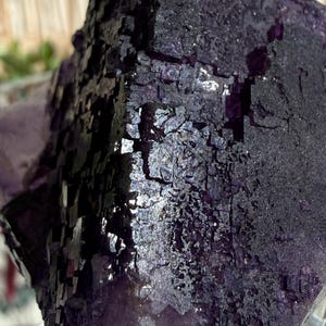 Puede incluir: Primer plano de un grupo de cristales de fluorita de color p&uacute;rpura oscuro. El cristal tiene una superficie rugosa y texturizada con una mezcla de tonos p&uacute;rpura oscuro y claro. La estructura cristalina es visible, con muchas formaciones peque&ntilde;as y cuadradas. El aspecto general es brillante y reflectante.