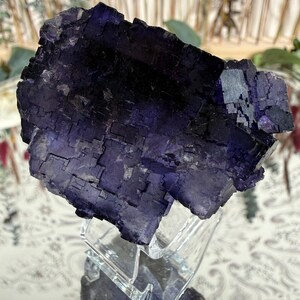 Puede incluir: Un gran grupo de cristales de fluorita de color p&uacute;rpura oscuro. El cristal tiene una estructura geom&eacute;trica en bloques con un color profundo y saturado. Se exhibe sobre un soporte de acr&iacute;lico transparente, reflejando la luz y mostrando su belleza natural. El fondo est&aacute; borroso.