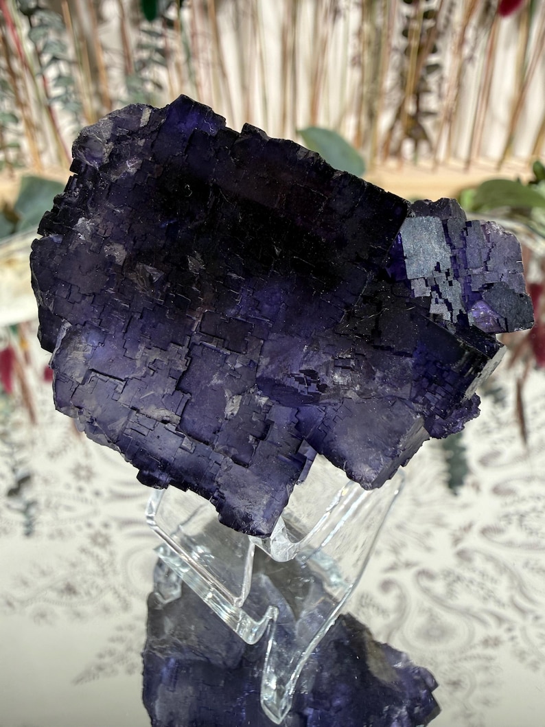Puede incluir: Un gran grupo de cristales de fluorita de color p&uacute;rpura oscuro. El cristal tiene una estructura geom&eacute;trica en bloques con un color profundo y saturado. Se exhibe sobre un soporte de acr&iacute;lico transparente, reflejando la luz y mostrando su belleza natural. El fondo est&aacute; borroso.
