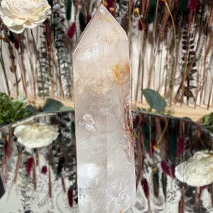 May include: A clear quartz crystal point with internal inclusions of orange and brown hues. The crystal is standing upright on a reflective surface, with dried flowers and decorative elements in the background. The crystal is approximately 6 inches tall.
