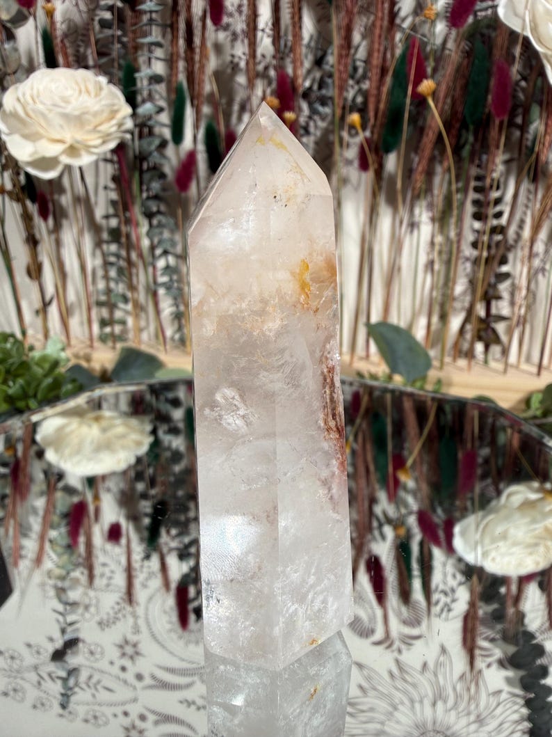 May include: A clear quartz crystal point with internal inclusions of orange and brown hues. The crystal is standing upright on a reflective surface, with dried flowers and decorative elements in the background. The crystal is approximately 6 inches tall.