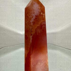 May include: A polished, tower-shaped crystal point featuring a vibrant gradient of deep burnt orange and translucent pale peach hues. The stone displays natural internal inclusions, varied mineral patterns, and a smooth, faceted surface.