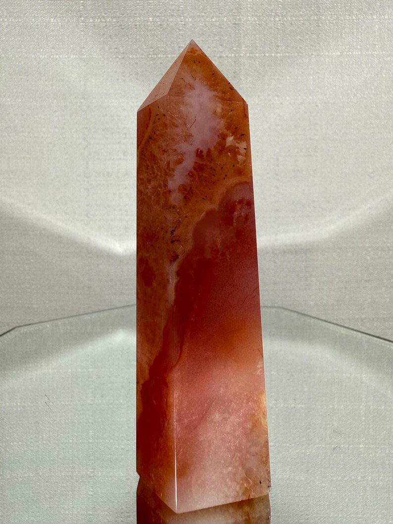 May include: A polished, tower-shaped crystal point featuring a vibrant gradient of deep burnt orange and translucent pale peach hues. The stone displays natural internal inclusions, varied mineral patterns, and a smooth, faceted surface.
