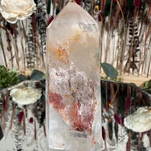 May include: A clear quartz crystal point with internal red and orange mineral inclusions. The crystal is faceted and stands upright, reflecting light. The background features dried flowers and decorative elements, suggesting a focus on natural decor and crystals.