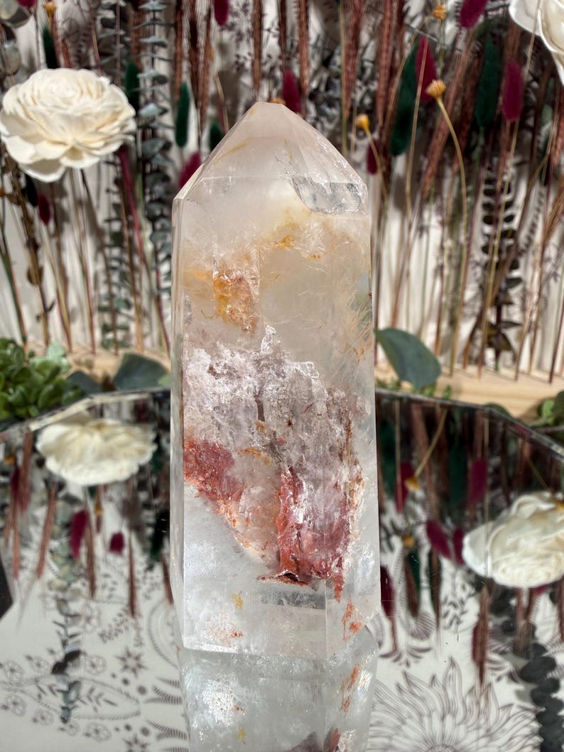 May include: A clear quartz crystal point with internal red and orange mineral inclusions. The crystal is faceted and stands upright, reflecting light. The background features dried flowers and decorative elements, suggesting a focus on natural decor and crystals.