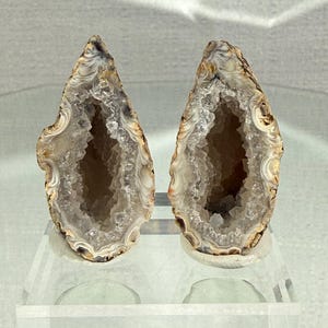 May include: Two teardrop-shaped agate geode halves standing upright. Each geode features a hollow center lined with sparkling, translucent quartz crystals, surrounded by concentric bands of white, tan, and brown mineral layers on the exterior.